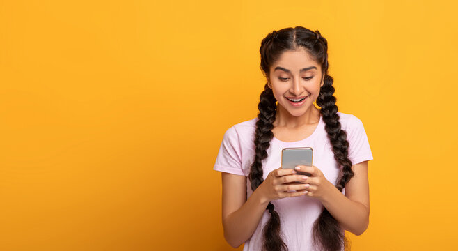 People And Technology Concept. Portrait of smiling cheerful young indian lady using her cellphone, chatting with friends online, standing isolated on yellow studio background, banner, free copy space - Powered by Adobe