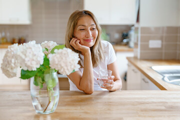 Relaxing moment at home with a woman enjoying a glass of water while contemplating in a bright kitchen surrounded by flowers