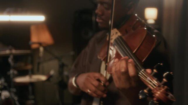 Rack focus shot of talented young African American man playing violin in studio and smiling enjoying music, focus flowing from face to hands