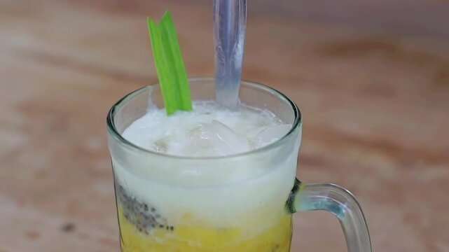 Refreshing yellow cendol drink with basil seeds, ice, and pandan garnish in a clear mug. Perfect for tropical beverage, traditional Asian dessert, and exotic culinary themes.