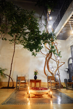 A cozy indoor courtyard with a tree, two chairs, a small table, and a decorative bowl with floating candles and flowers, creating a warm and inviting atmosphere. Moroccan Riad Villa.