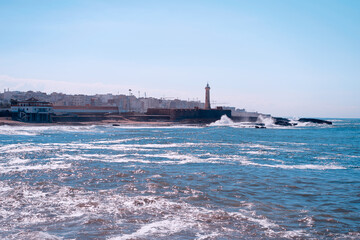 A beautiful coastal scene featuring a historic lighthouse against a backdrop of a vibrant town and ocean waves under a bright blue sky, creating a tranquil and scenic atmosphere.