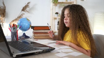 girl studying at desk with laptop and globe writing homework on card using pen while student organizes study material for school and education, child focused on study and homework task with book - Powered by Adobe