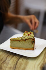 Close-up of a delicious slice of pistachio cheesecake served on a white plate. The dessert is topped with green frosting, chopped pistachios, and dried flower petals