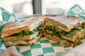 Freshly grilled sandwich with layers of meat, cheese, and greens in toasted bread on green checkered paper liner