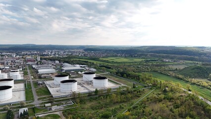 Drone view of industrial oil tanks and factory buildings beside green fields and distant hills. A striking contrast between technology and nature, industry and environment, progress and balance.