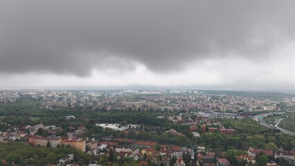 Aerial cityscape under dramatic storm cloud, showing urban districts, residential blocks, green areas, and highway. Dark sky creates moody atmosphere over the modern city.