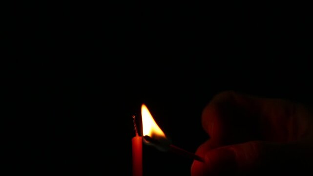 Woman lights a candle with a match on dark black background in holiday celebration. Candle burning fire flame with space. Magic ritual with twisting of candles. Close-up romantic emotion