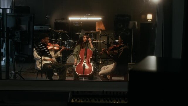 Wide angle shot of young string trio playing instruments in live room at recording studio, shot from behind glass window in producers booth, copy space