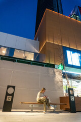 Urban Contemplation: A person sits on a bench, engrossed in their phone, amidst a backdrop of modern architectural marvels and twilight hues. A scene of serene solitude in the urban landscape.