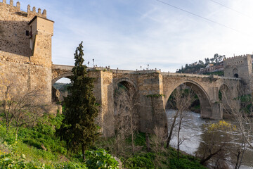 Toledo Bridge: Spain's historic San Martín Bridge, gracefully spanning the Tagus River, is steeped in history and culture, with medieval stone arches, towers, and a timeless design under a sunny sky