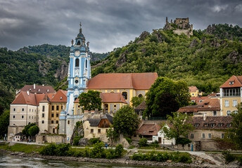 Image of a blue colored church steeple and red roofed buildings in a village along the banks of the Danube.