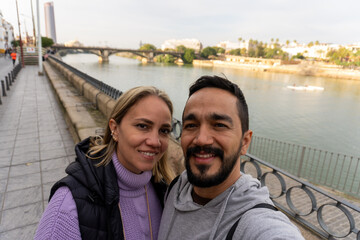 Selfie on a scenic bridge in Seville, Spain: a couple poses on a bridge in front of the camera, with a beautiful river and cityscape in the background.