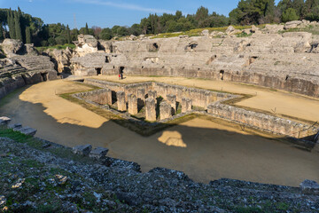 Ancient Roman Amphitheater itálica: A grand stone amphitheater stands proudly against a backdrop of verdant trees, the arena's walls weathered by time and history.