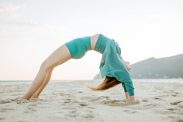 Young woman practicing yoga on the beach at sunrise, performing advanced backbend pose (Urdhva Dhanurasana). Outdoor workout by the ocean for strength, flexibility, and mindfulness
