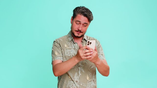 Middle-aged man scrolls smartphone casually, then suddenly hits forehead with palm realizing he forgot something important. Young adult guy isolated on blue background showing memory lapse and urgency