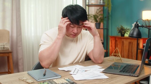 Asian man sitting at home table grips head, unpaid bills scattered with calculator and laptop, shoulders droop. Chinese guy sighs, showing financial stress and urgently seeking workable answer now.