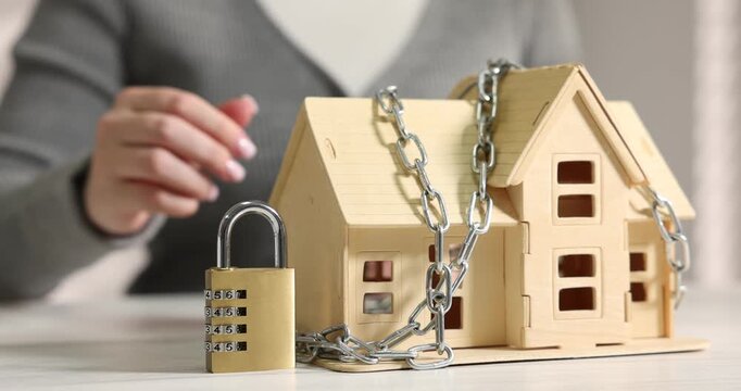 Home security. Woman putting padlock near house model with chain at white table, closeup