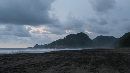 Dramatic seascape showing dark sand, cloudy skies, and misty hills along the shoreline during a moody, overcast day.	