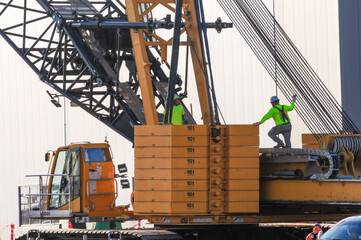 Construction workers secure rigging on a large crane at a stand-alone electric battery manufacturing plant under construction, with counterweights and lifting components visible on heavy equipment
