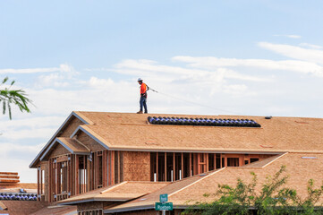 A construction worker walks along the roof of a wood-framed house under construction, with roofing tiles stacked nearby, indicating active residential building in a new housing development