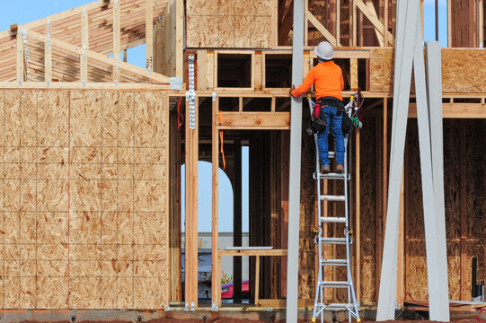 A construction worker wearing safety gear stands on a ladder while working on the wood framing of a new residential building, with structural sheathing and vertical panels at a home construction site