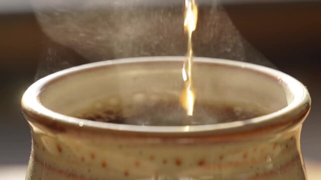 A stream of hot steaming black coffee being poured into a rustic handmade ceramic mug
