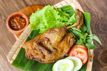 A serving of Bebek goreng (Indonesian fried duck) served on a banana leaf with fresh vegetables and a side of spicy sambal or chili sauce on a wooden table.