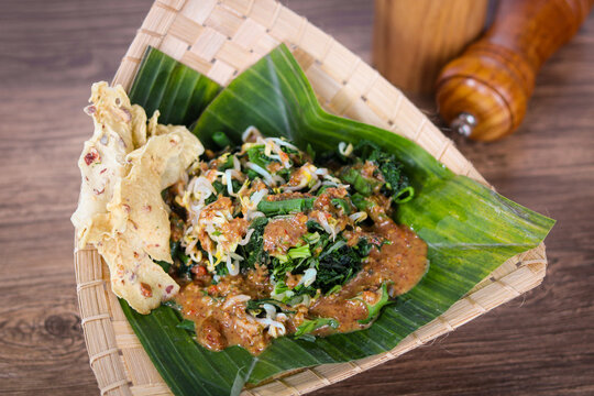 Pecel, a traditional Indonesian salad featuring blanched vegetables, bean sprouts, and long beans, drizzled with a peanut sauce. Served on a banana leaf with crispy rempeyek crackers.