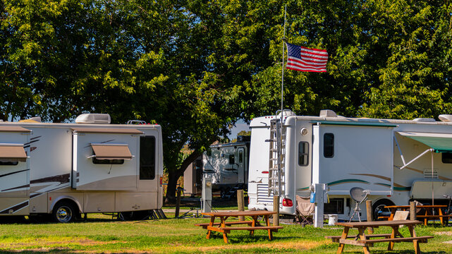 Several recreational vehicles are parked under a lush canopy of trees, creating a serene campground scene. An American flag flutters in the gentle breeze, while picnic tables invite visitors to relax.