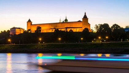 Obraz premium Illuminated castle on a hill overlooking a river at twilight, with a blurred boat in the foreground
