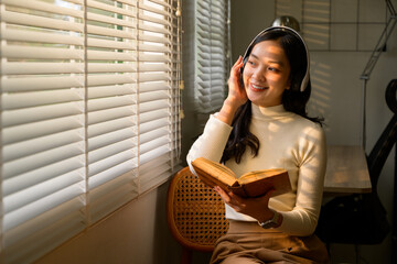 Young woman enjoying audiobook and reading book near window with blinds