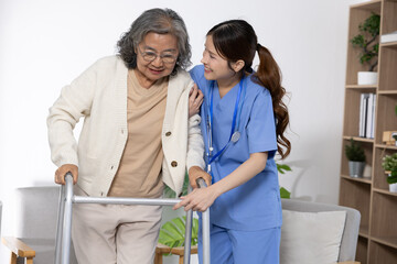 Asian Elderly woman using walker assisted by smiling female nurse in blue uniform providing supportive care in bright room with plants and bookshelf