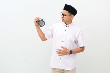 Asian Muslim man wearing koko shirt showing alarm clock, Ramadan fasting month concept. Holding stomach feeling hungry waiting for meal time