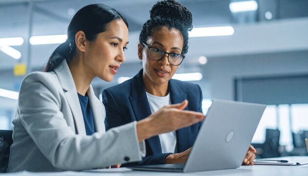 Close up confident Indian businesswoman mentor teaching African American intern. Using laptop, pointing at computer screen. Helping with corporate software. Detailed high quality image. 