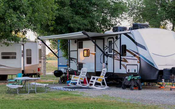 An inviting campsite features a modern RV with an extended awning, under which two white lounge chairs are positioned for relaxation. A picnic table and grill setup complete the cozy outdoor living 