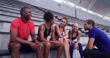 Fototapeta premium Conversation, break and exercise with friends on stadium bleachers for bonding or sports training. Fitness, recovery and rest with group of athlete people speaking in venue for workout discussion