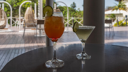 Two cocktail glasses on the table. The drinks are orange and green. Lime slices on the edges. The soft background is a terrace, resort furniture, wicker swing chairs. Cuba. Varadero.