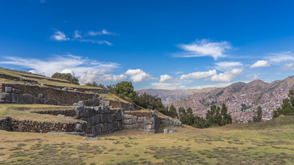 Ruins of the majestic ancient Inca fortress Saksaywaman. Terraces and tiers of walls made of polished large stones. Green grass on a meadow. In the distance, on a mountainside, the Cusco city