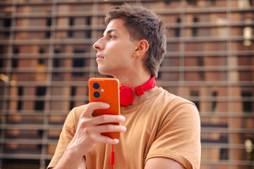 Side view of a young man wearing a brown t-shirt and red headphones around his neck, holding an...