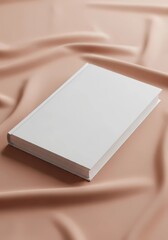 Fototapeta premium A closed white book rests on a smooth peach colored fabric surface