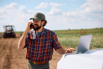 Happy farmer talking on mobile phone while working on laptop in the field. © Drazen