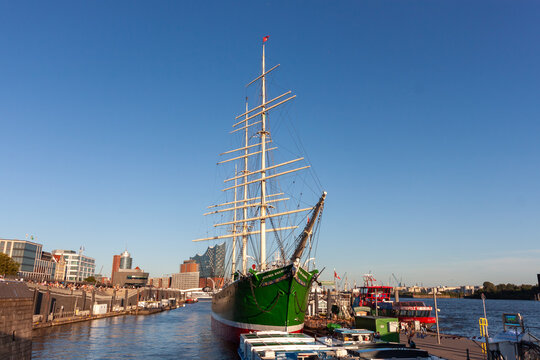 Panoramic view of the historical tall ship "Rickmer Rickmers" in Hamburg's harbor, with the modern Elbphilharmonie concert hall and cityscape visible in the background