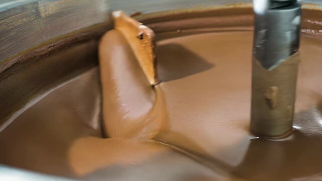 Industrial chocolate mixing in a stainless conche with central shaft and wooden scraper; macro view of glossy cocoa mass forming smooth folds and ribbons under continuous, controlled agitation.