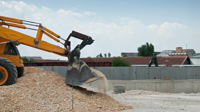 Yellow excavator unloading gravel at construction site, metal bucket pouring stones in mid‑air, dust and debris cascading, industrial fence and rooftops in background, wide daylight shot capturing mac