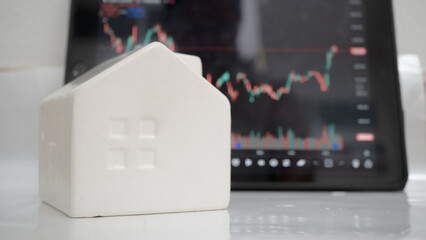 Small white ceramic house sits in front of tablet displaying stock market chart, symbolizing connection between real estate and financial markets