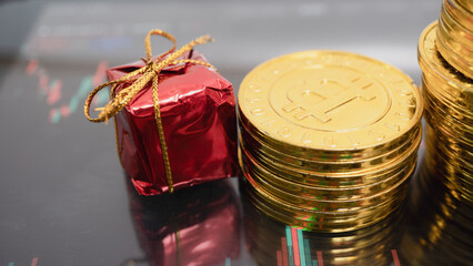 Small red gift box sits next to stacks of shiny gold coins with Bitcoin symbol, placed on digital financial chart background, symbolizing cryptocurrency investment and festive gifting