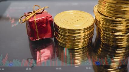 Stack of shiny gold coins and small red gift box with bow are placed on digital tablet displaying financial chart, symbolizing investment and holiday savings