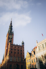 Fototapeta premium A low-angle view of the historic Town Hall in Gdansk, Poland, with its prominent clock tower reaching towards a bright sky. Other traditional buildings are visible nearby. 