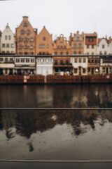 Traditional European buildings with colorful facades reflected in the water of a canal, shot through a fence with a blurred foreground.